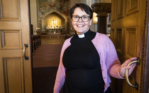 En kvinnlig präst i svart och med rosa kofta håller upp porten till hennes kyrka.