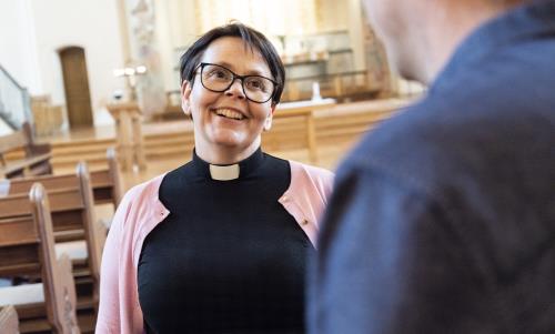 En kvinnlig präst i svart och med rosa kofta står och samtalar med en man i en ljus kyrka.