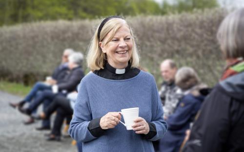 En kvinnlig präst står utomhus med en kaffemugg i handen och samtalar leende med någon.