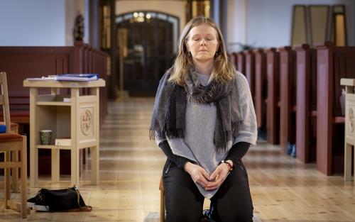 En kvinna sitter på en bönepall på en kyrkas golv, med knäppta händer och slutna ögon.