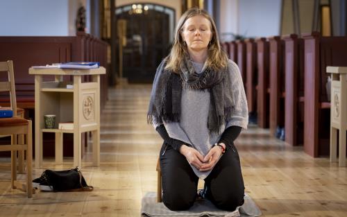 En kvinna sitter på en bönepall på en kyrkas golv, med knäppta händer och slutna ögon.