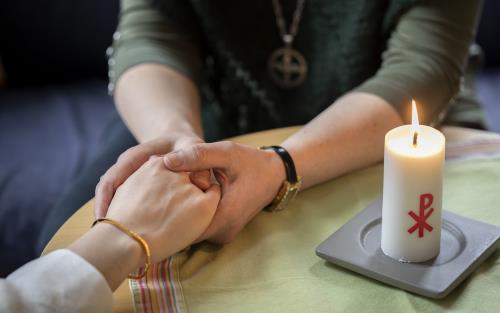 En diakon håller en annans hand i sina händer. Ett stearinljus med kristusmonogram brinner på bordet mellan dem.