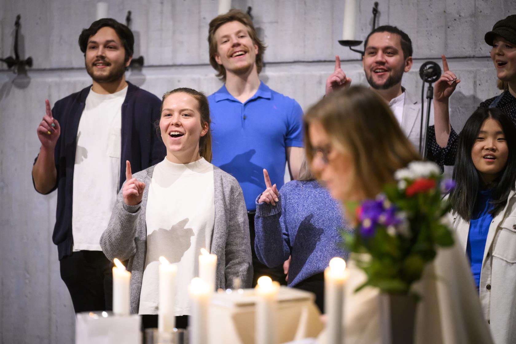 En blandad ungdomskör sjunger i en kyrka. De håller upp sina pekfingrar.