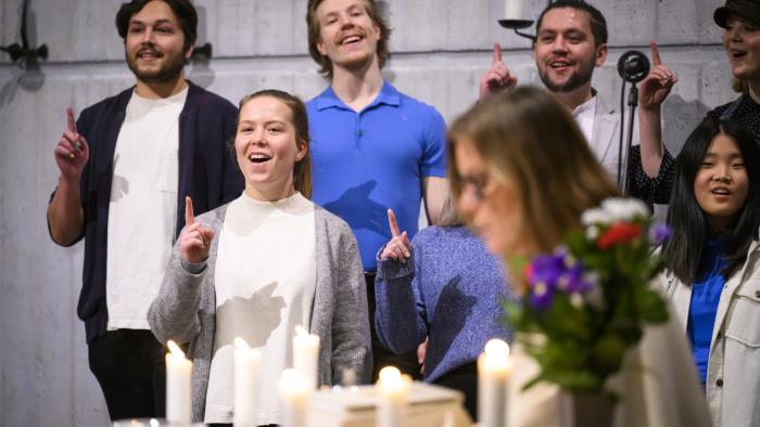 En blandad ungdomskör sjunger i en kyrka. De håller upp sina pekfingrar.