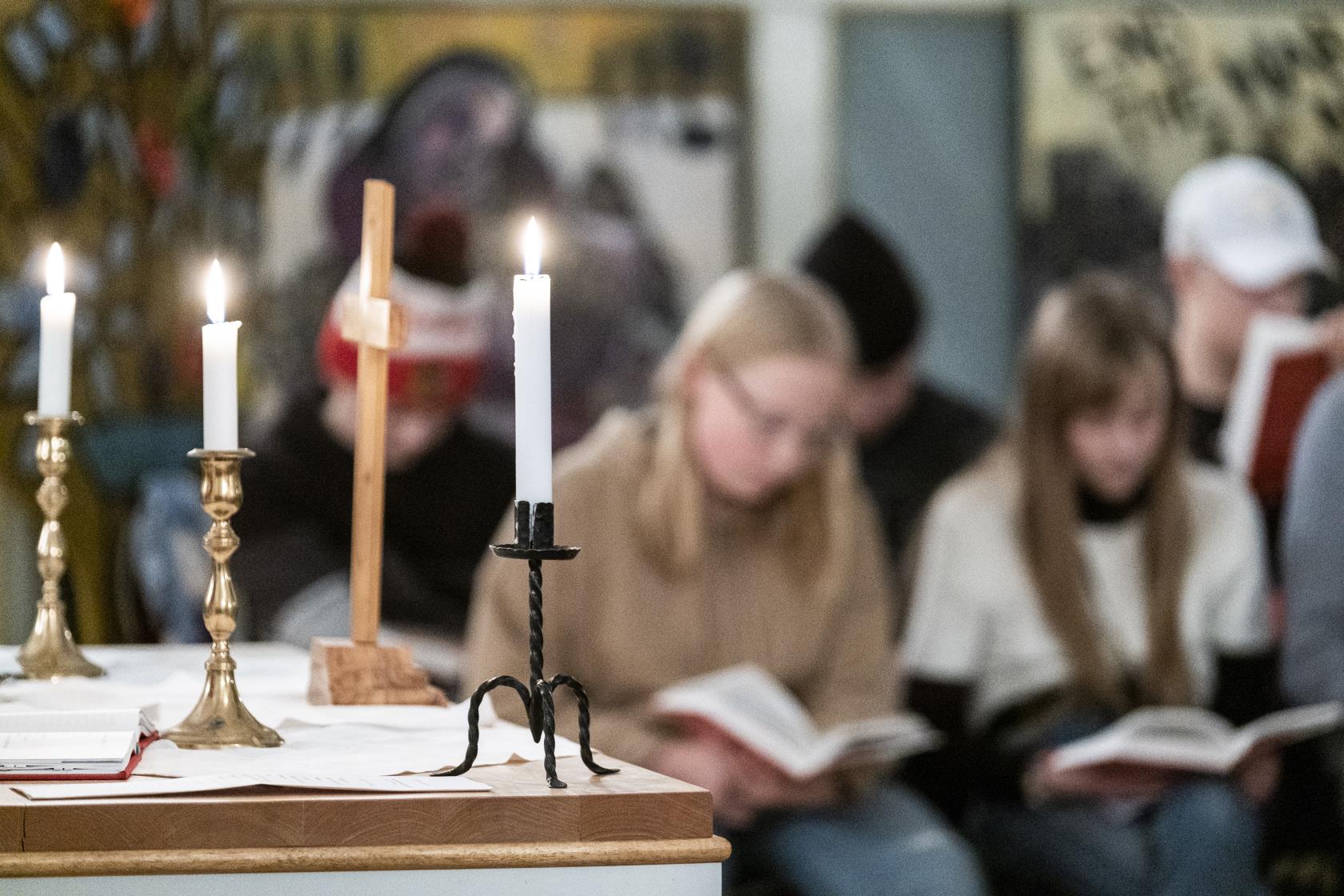 Ett litet träkors och tre ljusstakar med tända ljus står på ett altare. I bakgrunden syns suddigt en församling med uppslagna böcker.