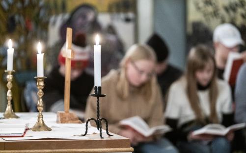 Ett litet träkors och tre ljusstakar med tända ljus står på ett altare. I bakgrunden syns suddigt en församling med uppslagna böcker.