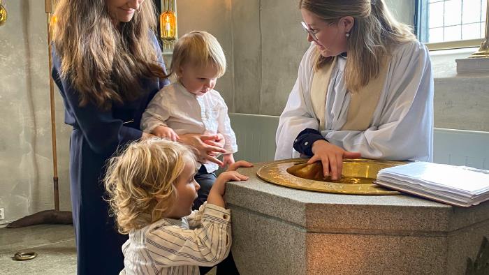 En kvinna och två små barn står tillsammans med en kvinnlig präst vid en dopfunt.