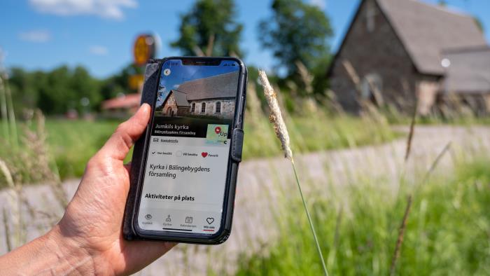 En person som kommit fram till en gammal kyrka håller en mobiltelefon i handen. På skärmen visas kyrkans namn och position på en karta.