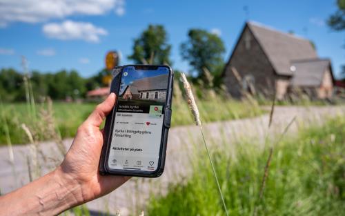 En hand håller i en mobil med skärmbild om kyrkguiden som visar adressen för kyrkan.