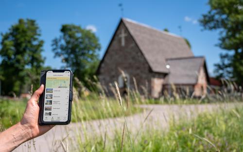 En person som kommit fram till en gammal kyrka håller en mobiltelefon i handen. På skärmen visas information om kyrkan.