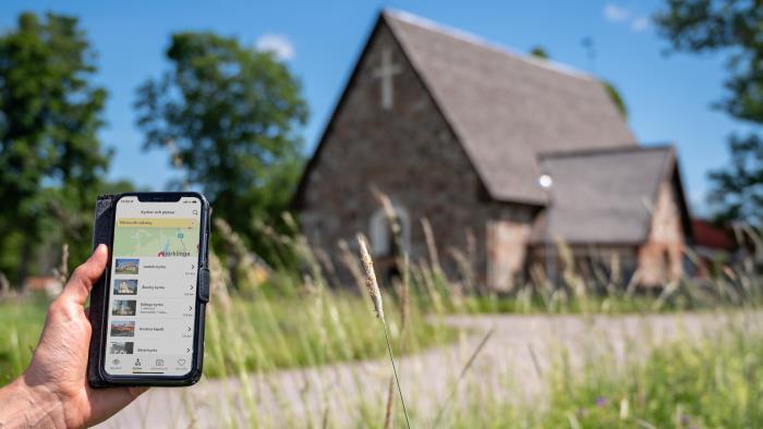 En person som kommit fram till en gammal kyrka håller en mobiltelefon i handen. På skärmen visas information om kyrkan.