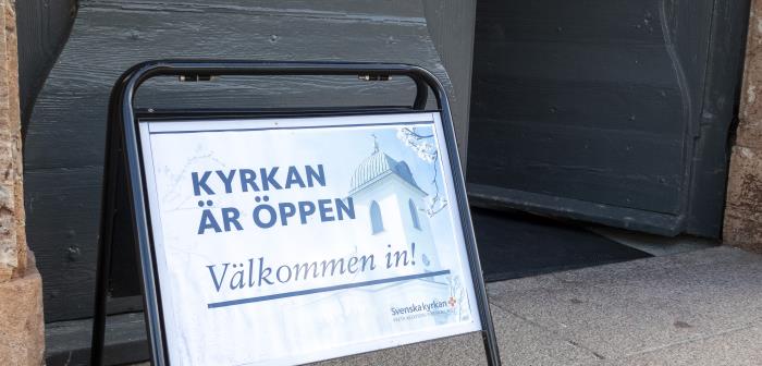 En skylt med texten Kyrkan är öppen välkommen in, står utanför en port.
