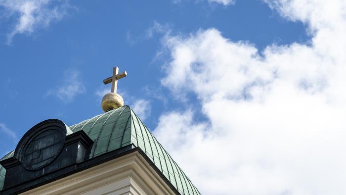Ett gyllene kors på ett kyrktak mot en blå och molnig himmel.