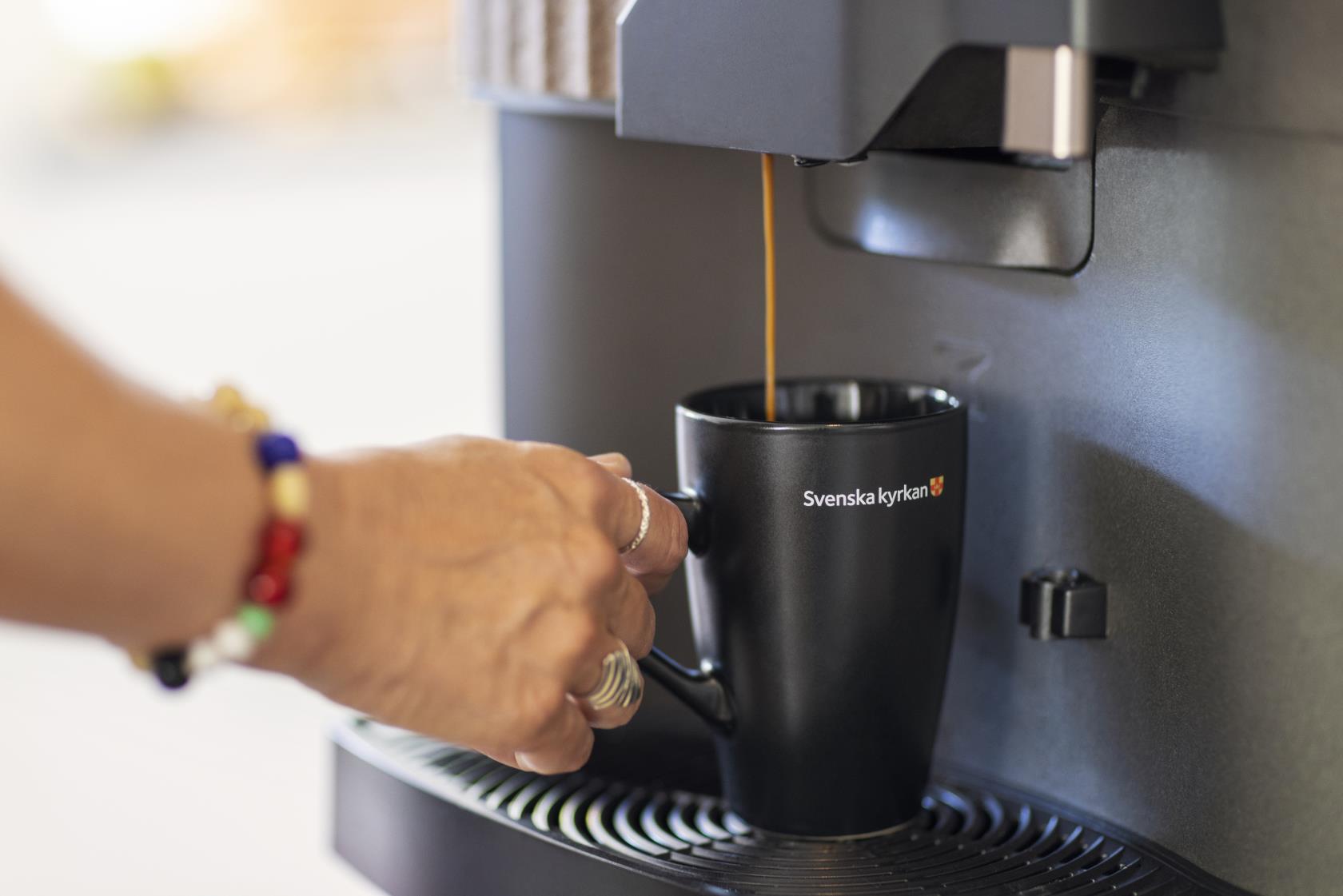 Någon tar kaffe från en kaffemaskin i en svart kopp med Svenska kyrkans logga.