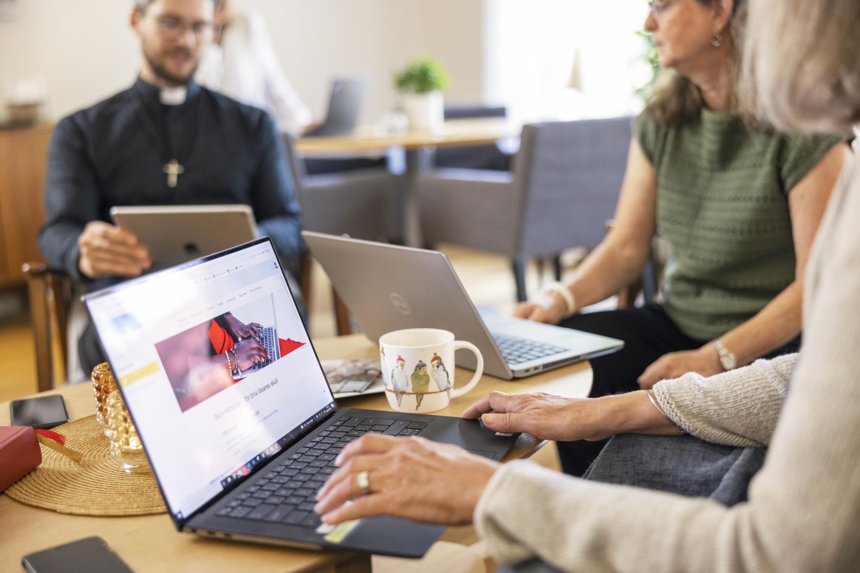 En manlig präst och två kvinnor sitter vid ett bord med varsin dator och samtalar.