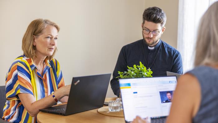 En manlig präst och några andra personer sitter runt ett bord med varsin laptop och samtalar.
