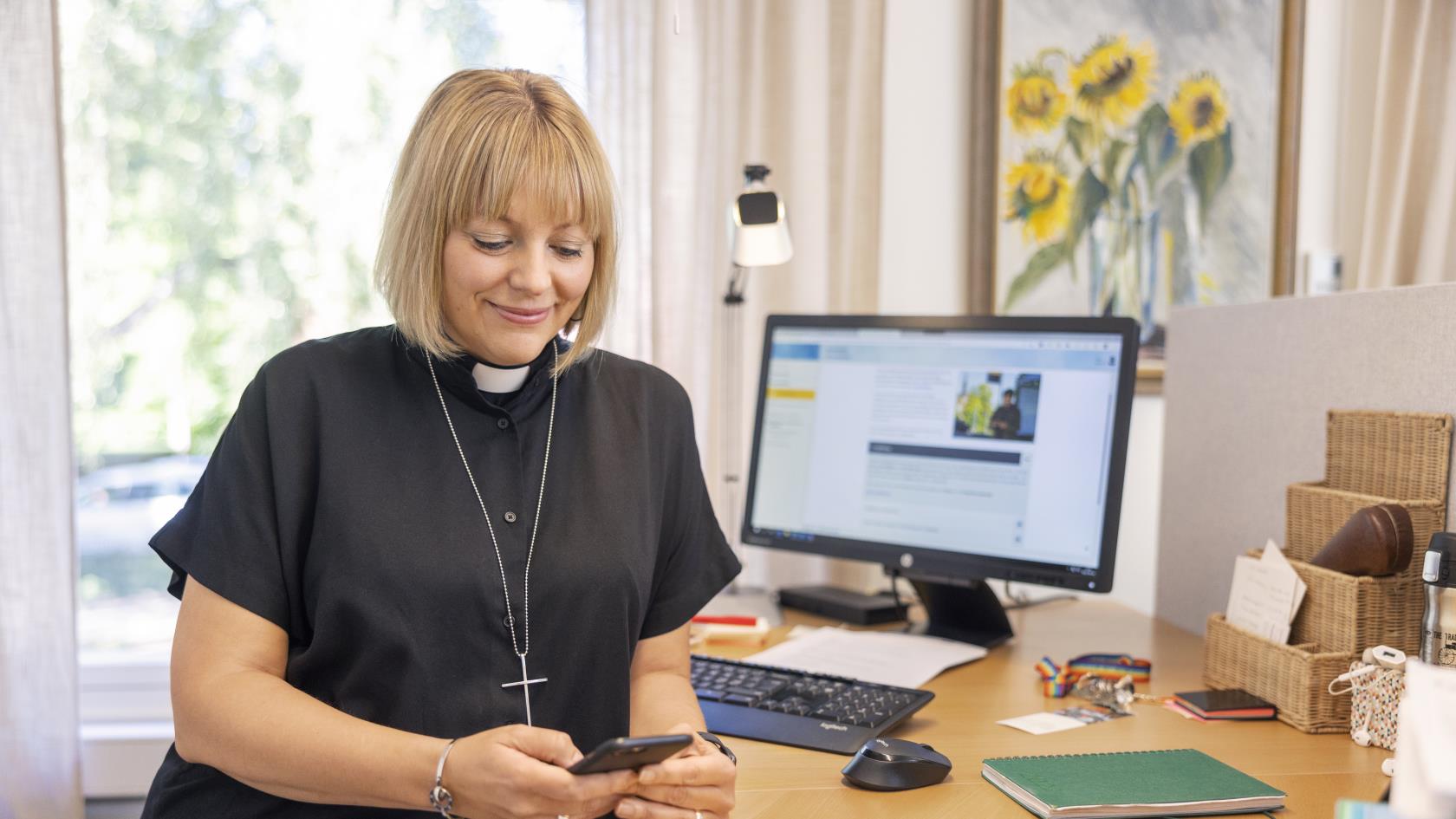 En kvinnlig präst står vid ett skrivbord med en mobiltelefon i handen.