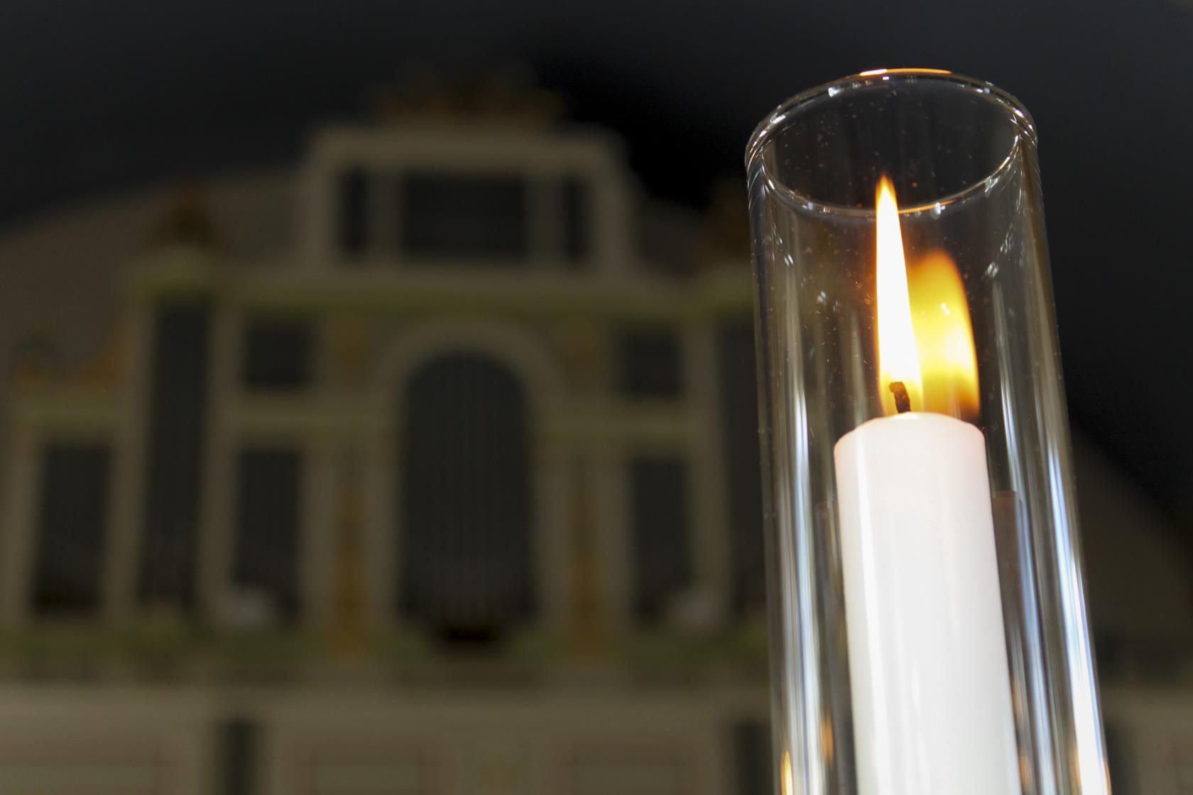 Närbild på ett brinnande ljus i en ljusstake med stormglas. En orgel i bakgrunden.