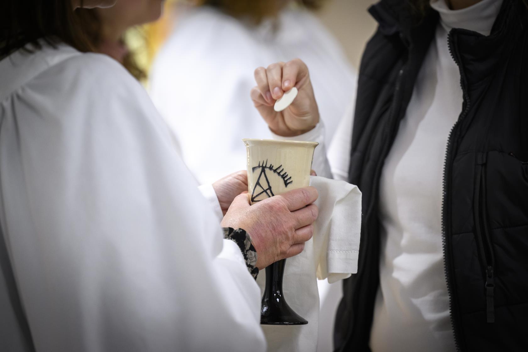 Någon i vit kåpa håller upp en kalk med nattvardsvin och en kyrkobesökare doppar oblaten i vinet.
