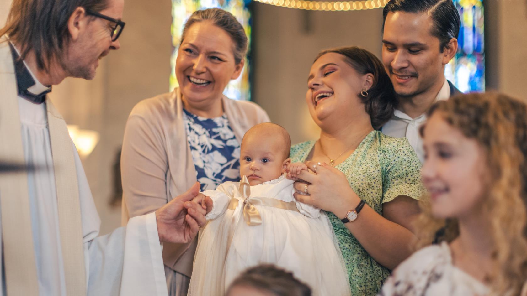 Ett dop i en kyrka. En kvinna med sin man bakom sig, håller barnet i famnen och skrattar. En manlig präst håller barnets hand.