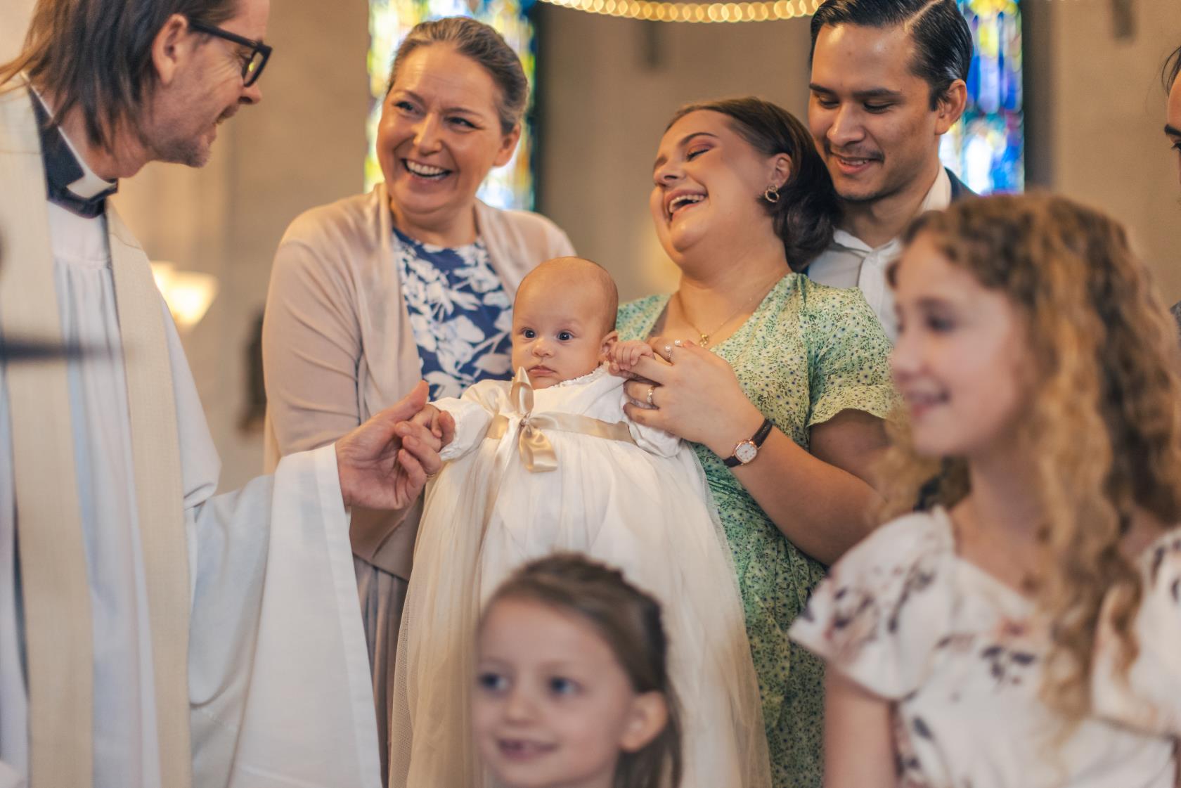 Ett dop i en kyrka. En kvinna med sin man bakom sig, håller barnet i famnen och skrattar. En manlig präst håller barnets hand.