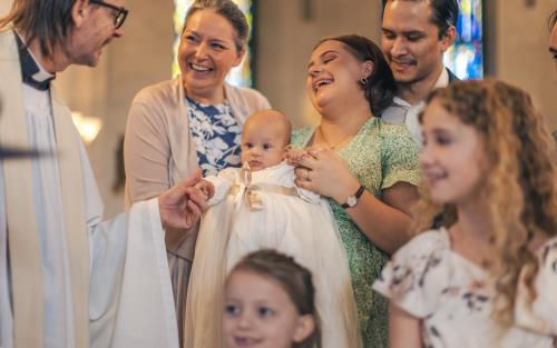 Ett dop i en kyrka. En kvinna med sin man bakom sig, håller barnet i famnen och skrattar. En manlig präst håller barnets hand.