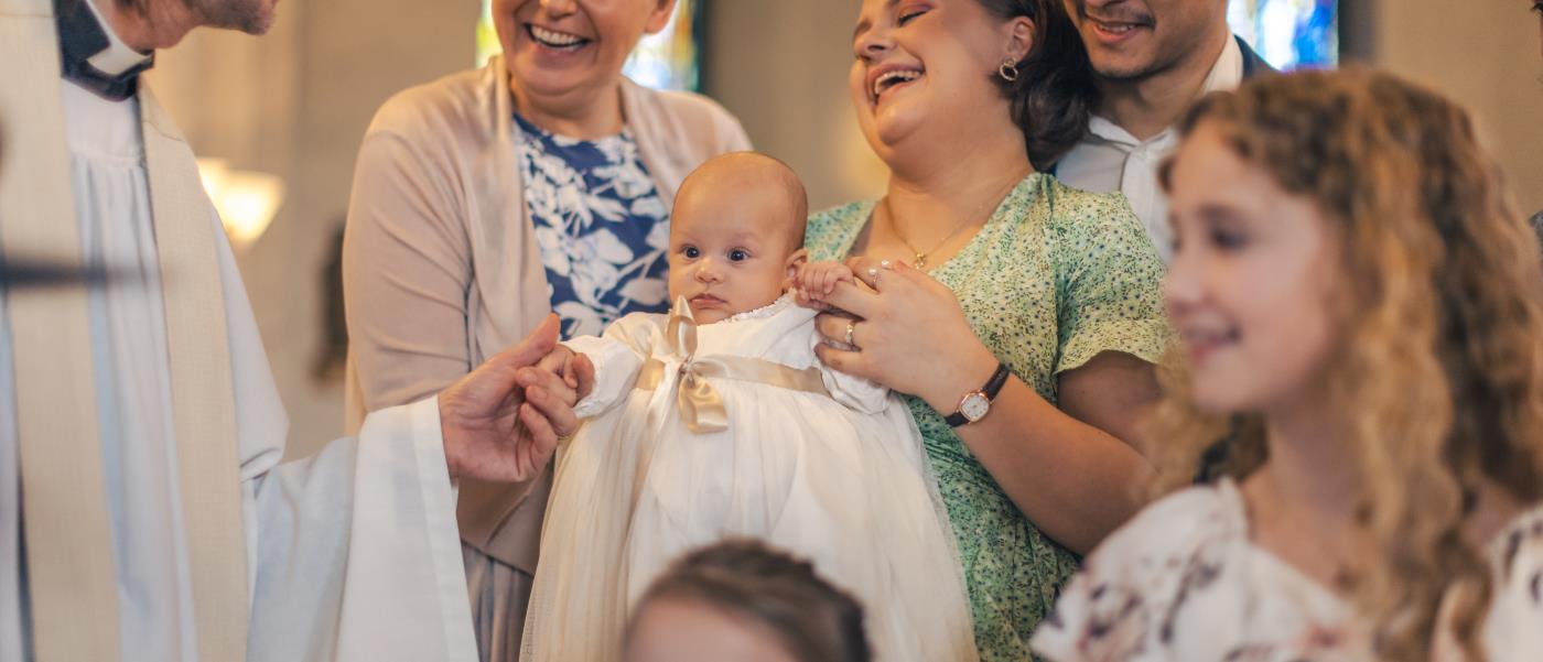 Ett dop i en kyrka. En kvinna med sin man bakom sig, håller barnet i famnen och skrattar. En manlig präst håller barnets hand.