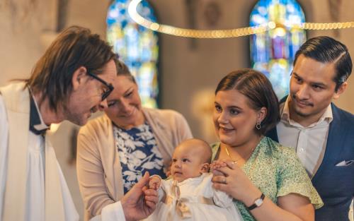 Ett dop i en kyrka. En kvinna med sin man bakom sig, håller barnet i famnen. En manlig präst håller barnets hand.