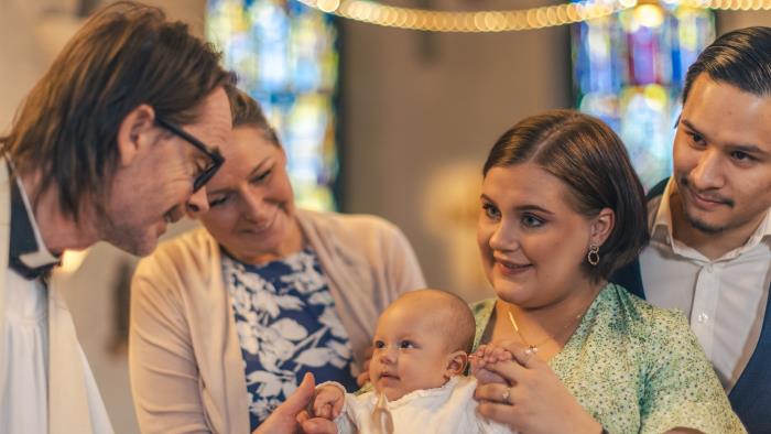 Ett dop i en kyrka. En kvinna med sin man bakom sig, håller barnet i famnen. En manlig präst håller barnets hand.