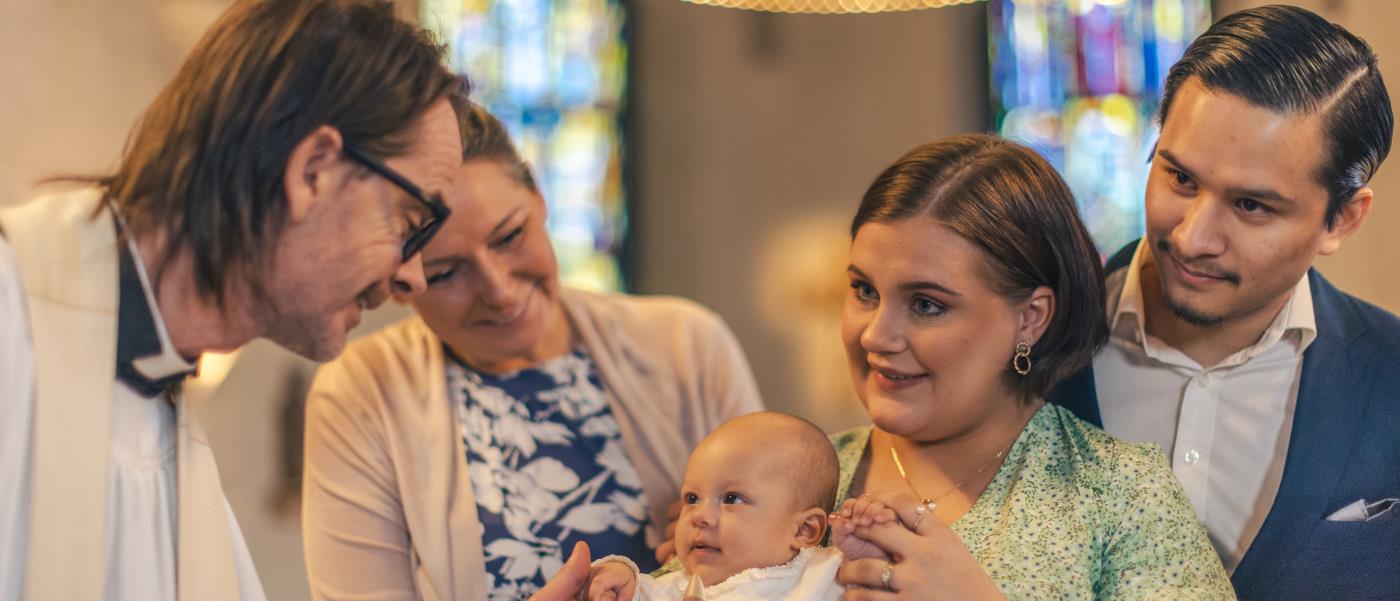 Ett dop i en kyrka. En kvinna med sin man bakom sig, håller barnet i famnen. En manlig präst håller barnets hand.