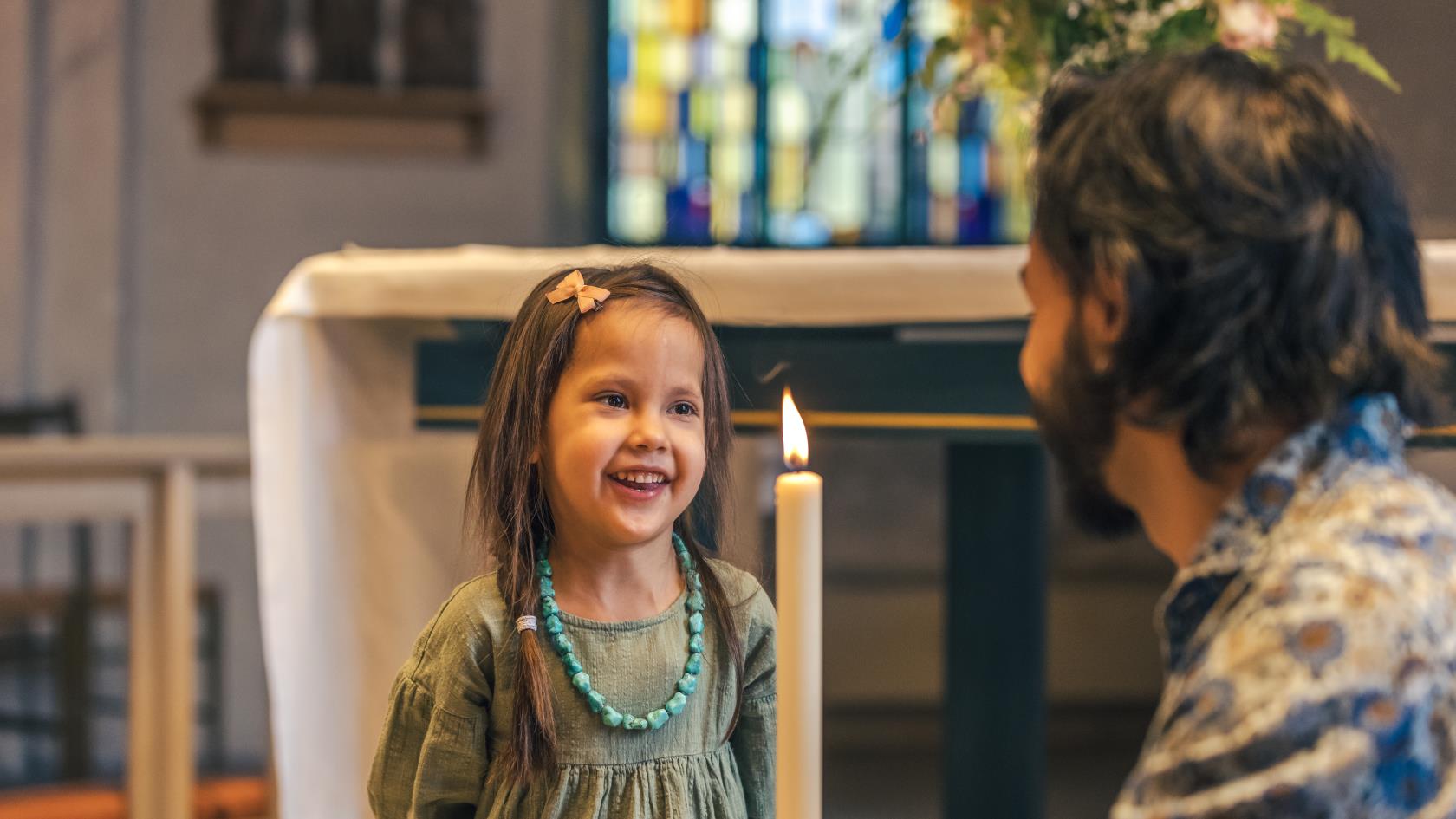 En man sitter på huk i en kyrka och håller i ett dopljus. En liten flicka står framför och ler stort.
