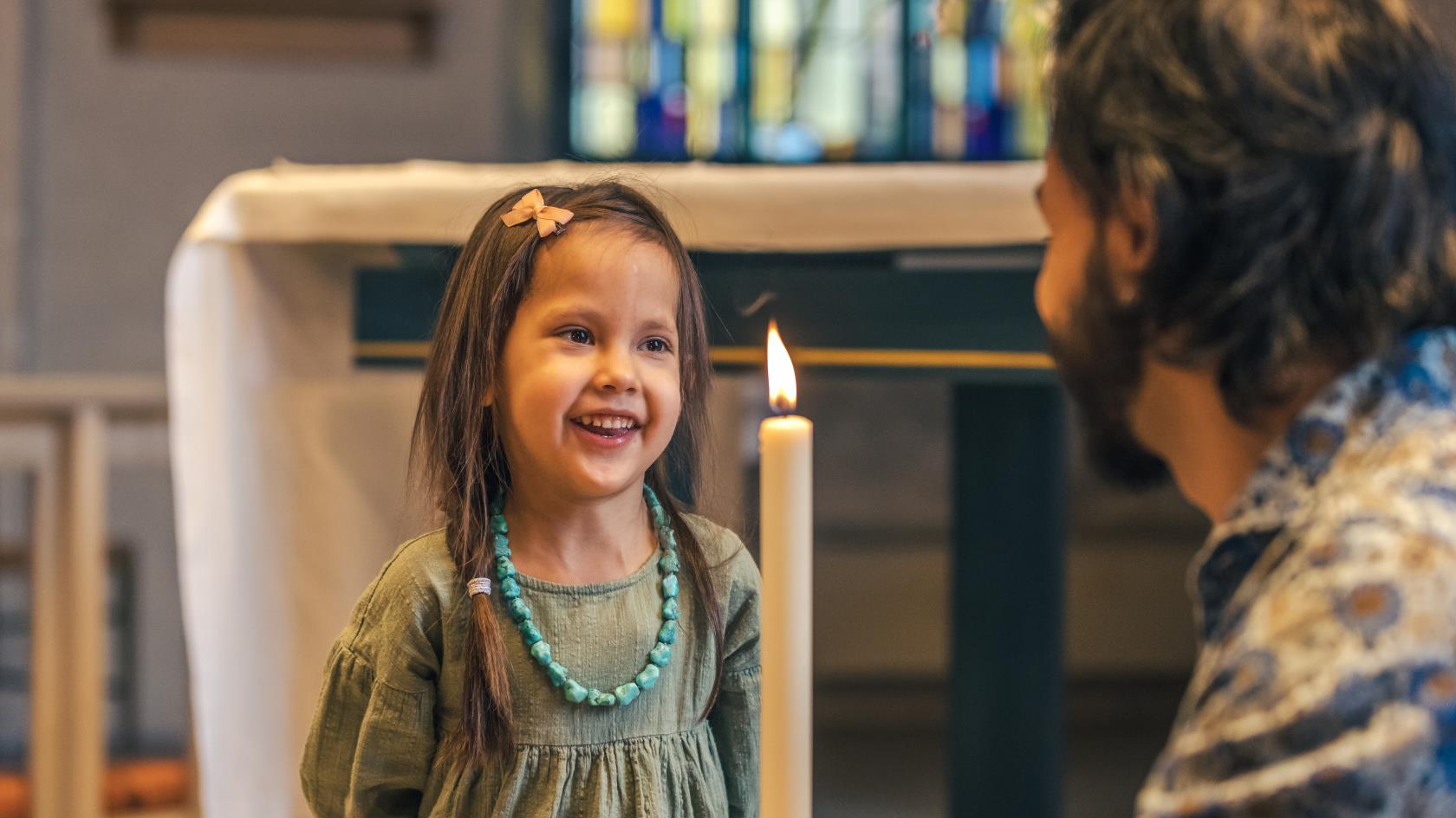 En man sitter på huk i en kyrka och håller i ett dopljus. En liten flicka står framför och ler stort.