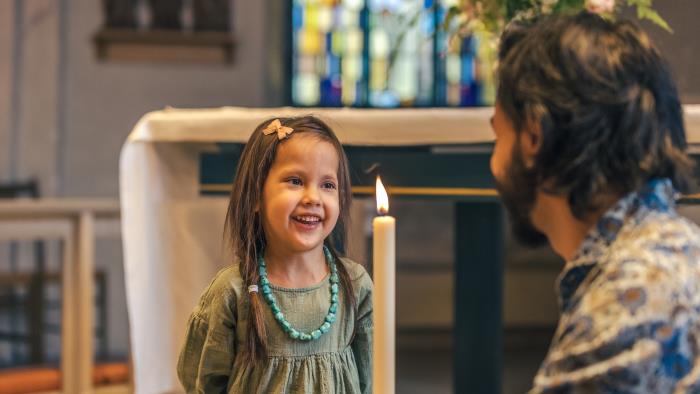 En man sitter på huk i en kyrka och håller i ett dopljus. En liten flicka står framför och ler stort.