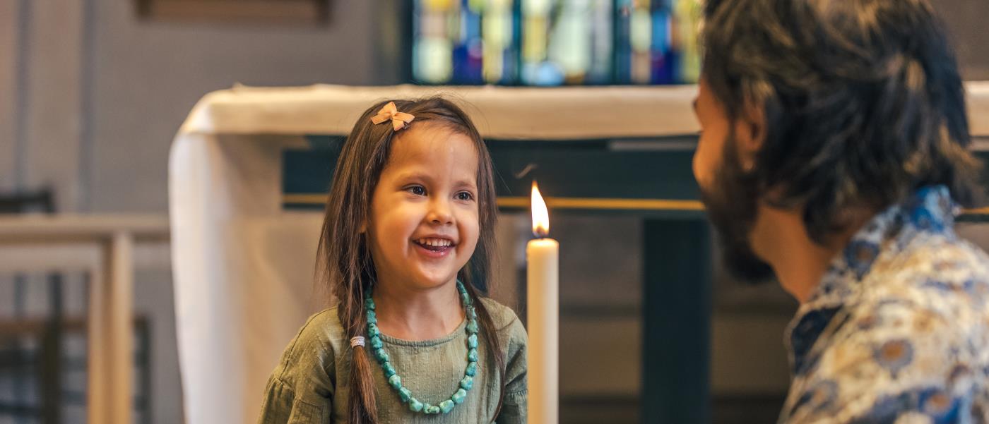 En man sitter på huk i en kyrka och håller i ett dopljus. En liten flicka står framför och ler stort.