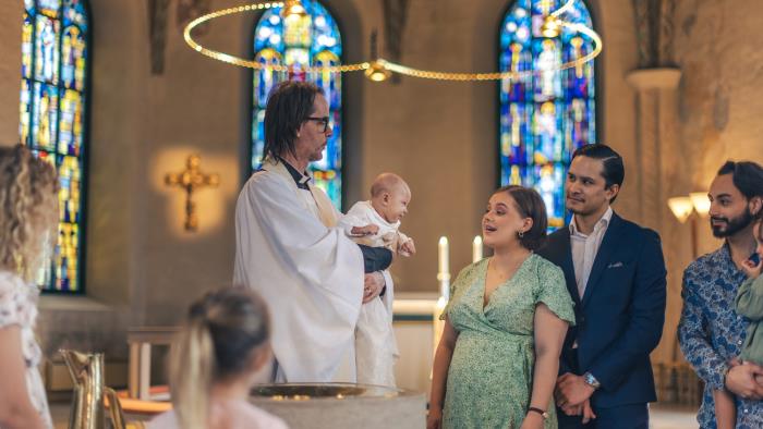 Ett dop i en kyrka. En kvinna och hennes man står bredvid prästen som håller barnet i famnen.