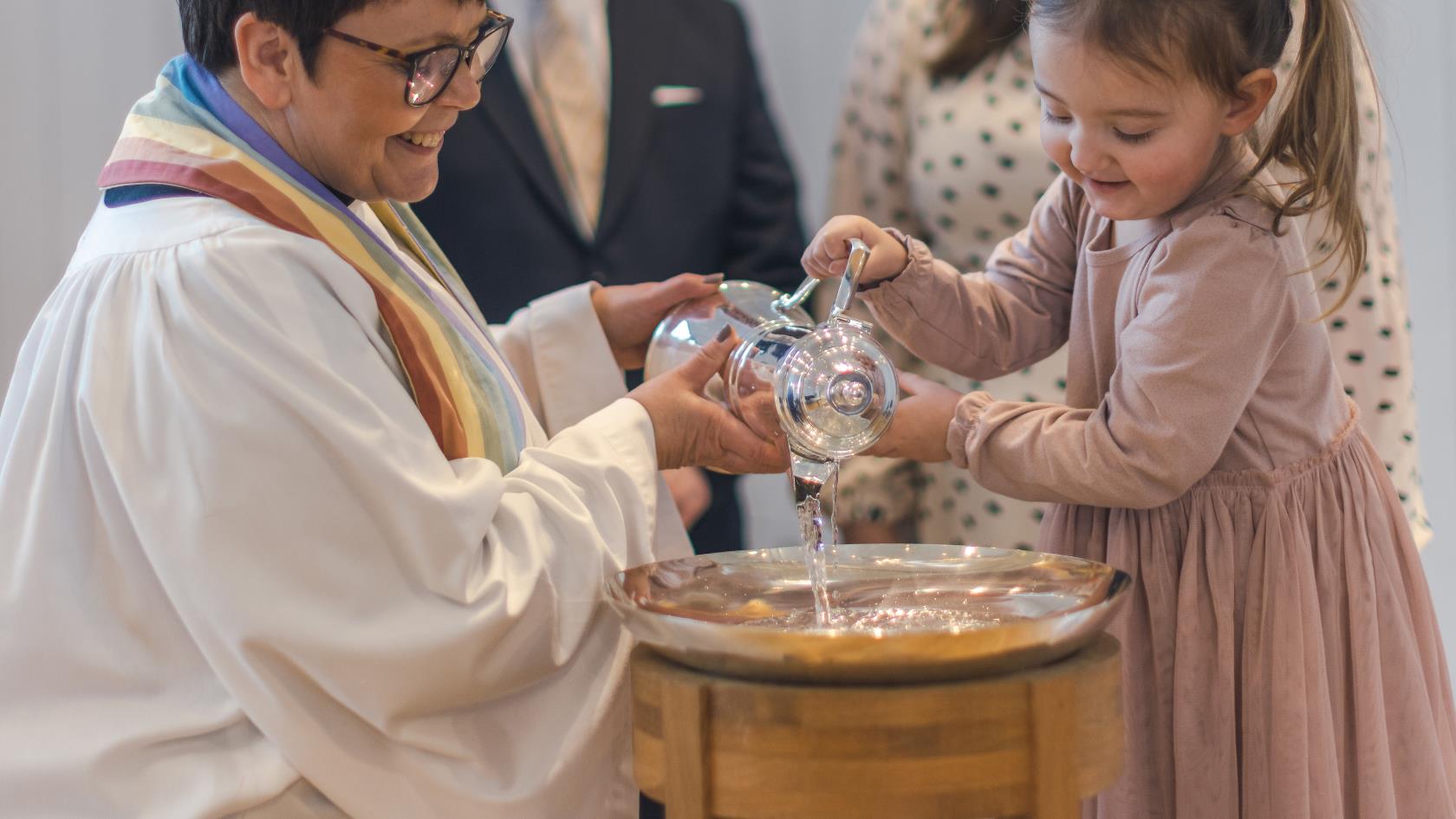 En kvinnlig präst och en liten flicka häller tillsammans vatten i en dopfunt. Två personer står bakom.
