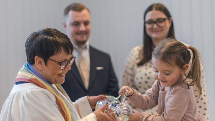 En kvinnlig präst och en liten flicka häller tillsammans vatten i en dopfunt. Två personer står bakom.