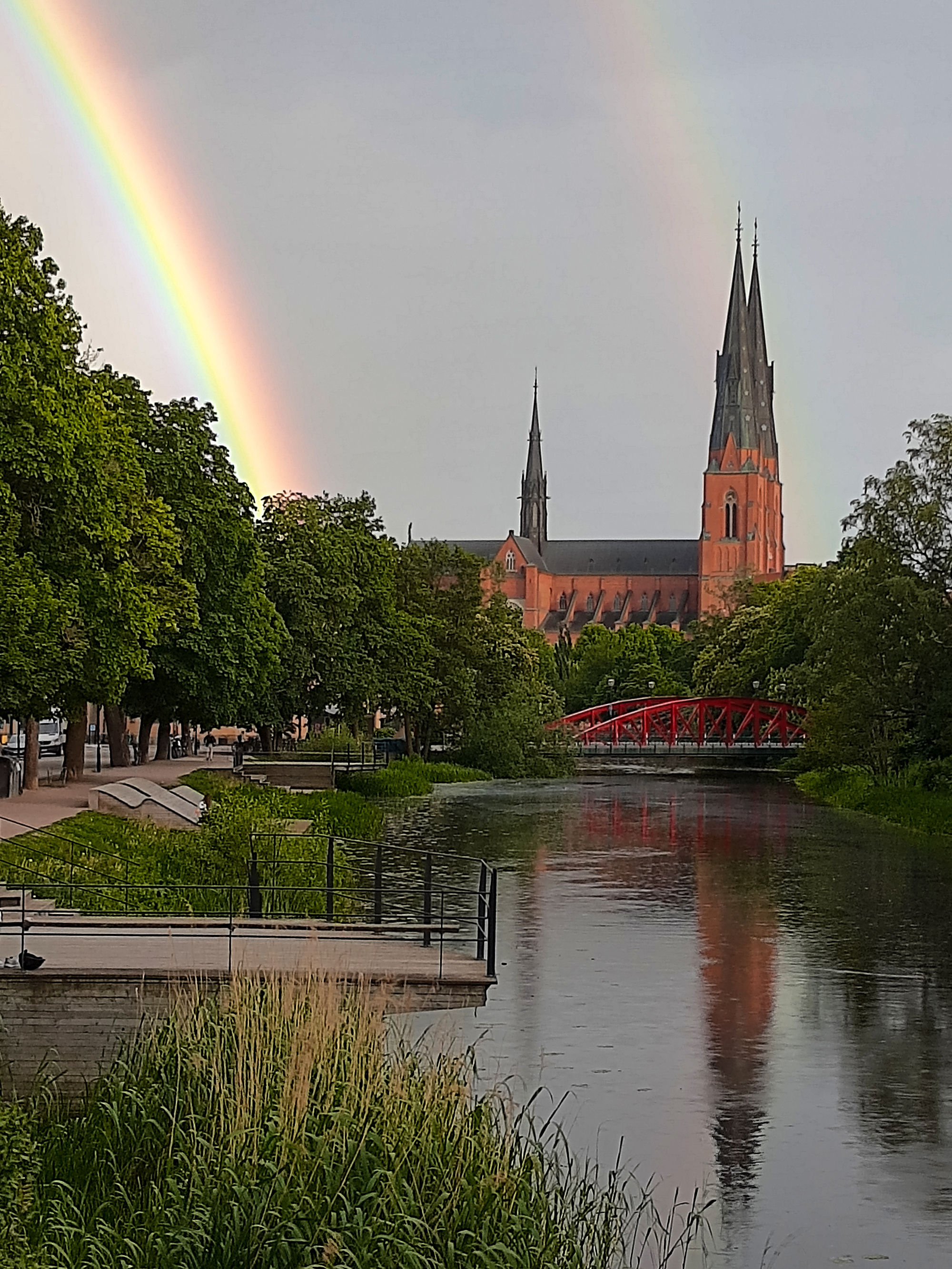 Dubbla regnbågar syns över Uppsala domkyrka. Kyrkan reflekteras i Fyrisån.