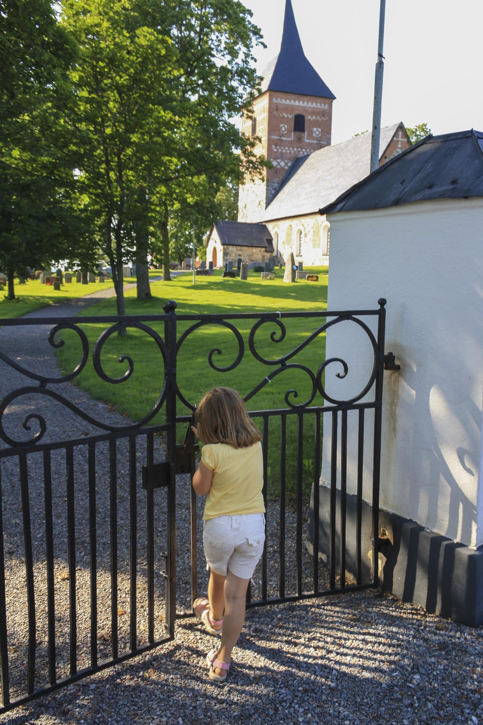 Ett litet barn öppnar järngrinden in till en kyrkogård med en kyrka i sten och rött tegel.
