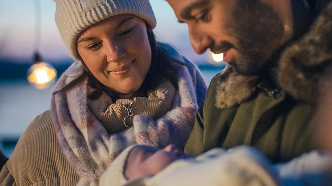 En man och en kvinna i ytterkläder står utomhus. Mannen håller i en bebis.