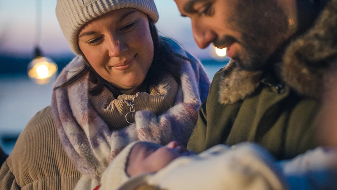 En man och en kvinna i ytterkläder står utomhus. Mannen håller i en bebis.