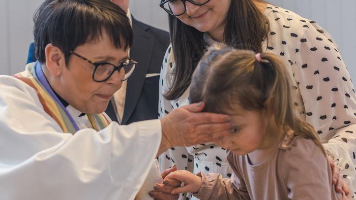 En kvinnlig präst skvätter dopvatten på en liten flickas panna. Föräldrarna står bakom.