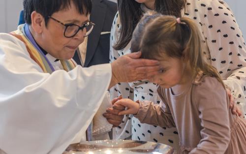 En kvinnlig präst skvätter dopvatten på en liten flickas panna. Föräldrarna står bakom.