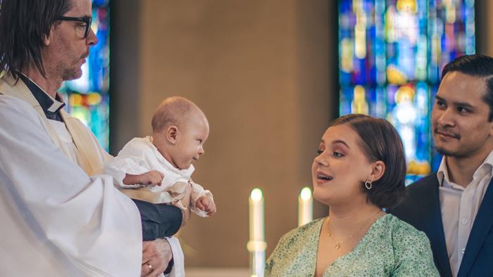 Ett dop i en kyrka. En kvinna och hennes man står bredvid prästen som håller barnet i famnen.