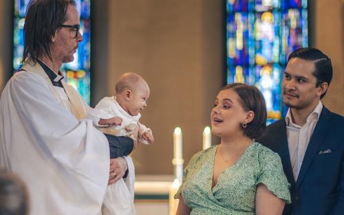 Ett dop i en kyrka. En kvinna och hennes man står bredvid prästen som håller barnet i famnen.