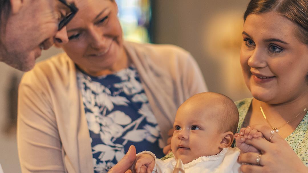 Ett dop i en kyrka. En kvinna med sin man bakom sig, håller barnet i famnen. En manlig präst håller barnets hand.