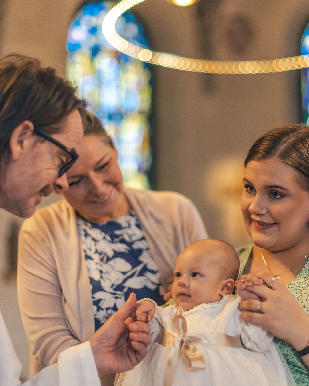 Ett dop i en kyrka. En kvinna med sin man bakom sig, håller barnet i famnen. En manlig präst håller barnets hand.