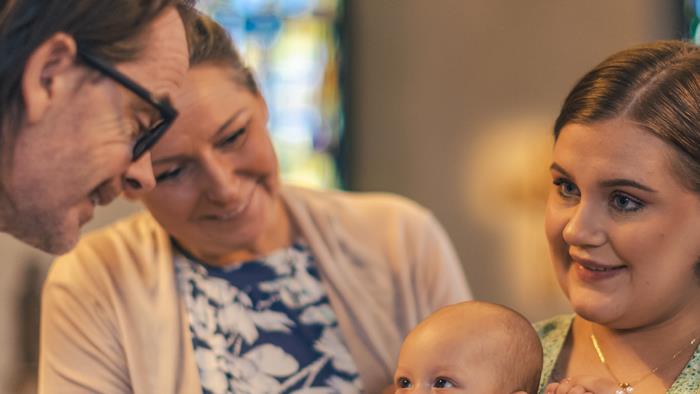 Ett dop i en kyrka. En kvinna med sin man bakom sig, håller barnet i famnen. En manlig präst håller barnets hand.