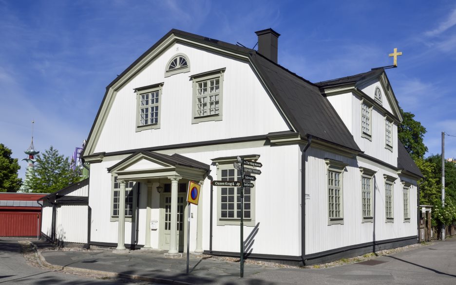 En stor vit trävilla med brutet tak av svart plåt. Från ena nocken hänger ett guldfärgat kors.