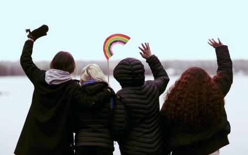 Fyra ungdomar står med ryggen mot kameran, vända mot vattnet och vinkar. En av dem håller i en regnbåge på en pinne.